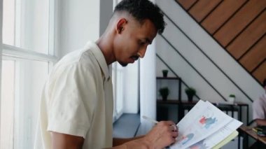 Genç siyahi bir adam evrak okuyor ve süslü bir ofiste kameraya bakıyor. Belgeler ve grafikler üzerinde çalışan bir adam. Verimli bir iş. Yüksek kalite 4k görüntü