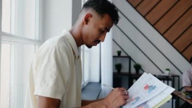 Genç siyahi bir adam evrak okuyor ve süslü bir ofiste kameraya bakıyor. Belgeler ve grafikler üzerinde çalışan bir adam. Verimli bir iş. Yüksek kalite 4k görüntü