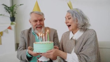 Yaşlı bir çift pastaya mum üflüyor ve doğum günü yemeğinde gülüyor. Pastel renkli parti. Mutlu büyükanne ve büyükbaba. Yüksek kalite 4k görüntü