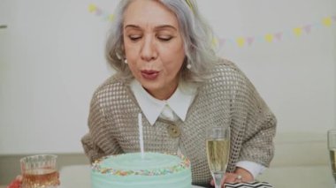 Yaşlı bir kadın doğum günü yemeğinde pastaya mum üflüyor. Pastel renkli parti. Mutlu büyükanne. Aile kucaklaşması ve büyükannenin doğum gününü kutlar. Yüksek kalite 4k görüntü
