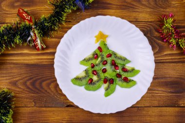 Kivi dilimleri ve nar ağacından yapılmış Noel ağacı ve ahşap masa üzerindeki Noel süsü. Üst Manzara. Noel ve Yeni Yıl bayramı tatlıları için yaratıcı bir fikir. Çocuklar için komik yemek fikri.