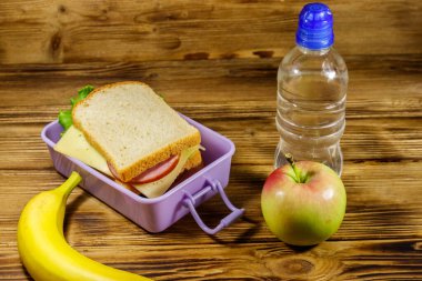 Sandviçli beslenme çantası, bir şişe su, muz ve elma ahşap bir masada.