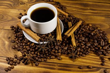 Bir fincan kahve, kavrulmuş kahve çekirdekleri, yıldız anason ve ahşap masada tarçın çubukları.