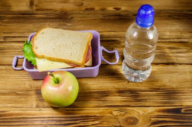 Sandviçli beslenme çantası, bir şişe su ve ahşap masada elma.