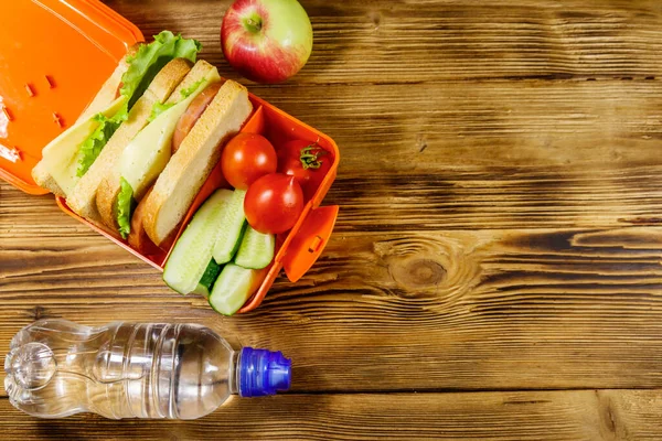 Bottle of water, apple and lunch box with sandwiches and fresh vegetables on a wooden table. Top view, copy space