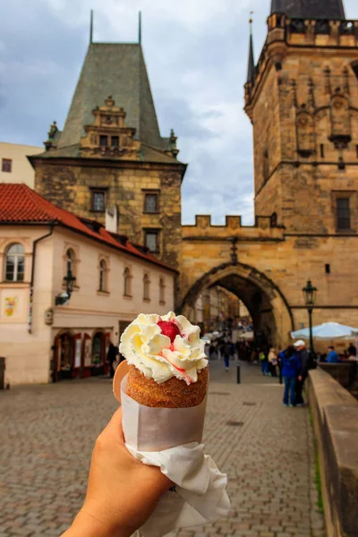 Female hand holding traditional czech cake trdelnik with whipped cream on the background of famous Charles bridge in Prague, Czech Republic