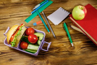 Okul konseptine geri dönelim. Okul gereçleri, kitaplar, elma ve beslenme çantası. Hamburgerler ve taze sebzeler ahşap bir masada.