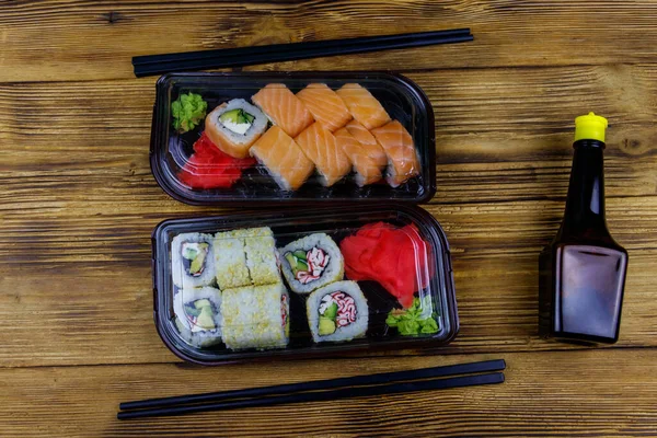 Set of sushi rolls in plastic boxes, soy sauce and chopsticks on wooden table. Top view. Sushi for take away or delivery of sushi in plastic containers