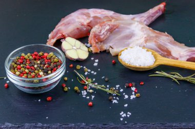 Çiğ tavşan bacağı ve siyah bir tahta üzerinde baharatlar.