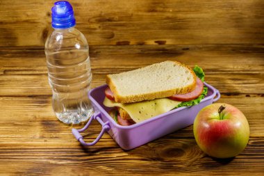 Sandviçli beslenme çantası, bir şişe su ve ahşap masada elma.