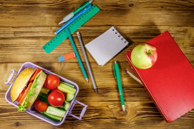 Okul konseptine geri dönelim. Okul gereçleri, kitaplar, elma ve beslenme çantası içinde hamburgerler ve ahşap bir masada taze sebzeler. Üst görünüm