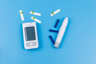 Dijital glukometre, tükenmez kalem, tek kullanımlık iğneler ve pastel mavi arka planda test şeritleri. Üst Manzara. Diyabet kavramı