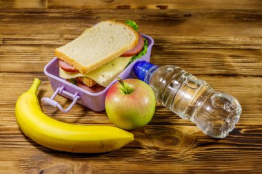 Sandviçli beslenme çantası, bir şişe su, muz ve elma ahşap bir masada.