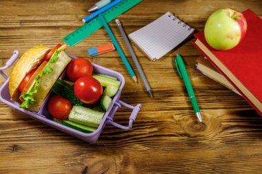 Okul konseptine geri dönelim. Okul gereçleri, kitaplar, elma ve beslenme çantası. Hamburgerler ve taze sebzeler ahşap bir masada.