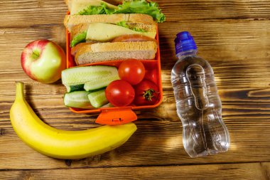 Bir şişe su, elma, muz, sandviç ve taze sebzelerle birlikte ahşap bir masada. Üst görünüm