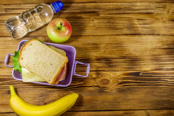 Sandviçli beslenme çantası, bir şişe su, muz ve elma ahşap bir masada. Üst görünüm, alanı kopyala