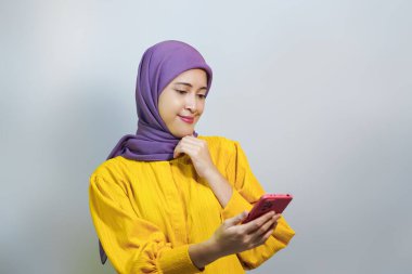 Heyecanlı genç Asyalı Müslüman kadın tesettür ve sarı elbise giyiyor cep telefonu kullanıyor ve mutlu görünüyor, beyaz stüdyo arka planında iyi haberler alıyor.