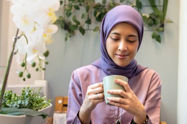 Tesettürlü kadın balkonda otururken bardağına bakıyor.