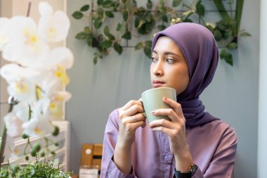 Başörtülü güzel bir kadın elinde yeşil bir ordu camı tutarak balkonda oturuyor. Önüne bakıyor, hayal kuruyor ya da düşünüyor.