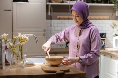 Mor bir elbise giyen güzel Asyalı kadın kolye ışığı ve lavabonun yanında modern bir mutfakta hamur yapıyor.