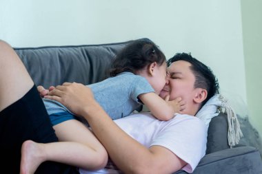 Asian man lying on sofa in living room with little girl on top, both smiling, father and daughter relationship. clipart