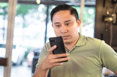 Endonezyalı bir adam kapalı bir kafede oturuyor, cep telefonuna bakıyor ya da telefonda gülümsüyor, telefona gülümsüyor, yeşil polo tişört giyiyor..