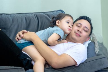 Asian man lying on sofa in living room with little girl on top, both smiling, father and daughter relationship. clipart