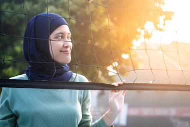 Muslim woman in hijab with brown eyes holding volleyball net in morning sunlight. clipart