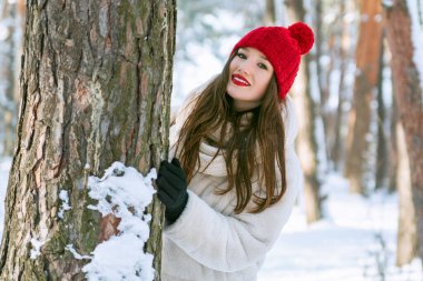 Kozalaklı bir kış ormanında ağaçların arkasında duran mutlu, esmer, beyaz bir kadının portresi. Açık havada.