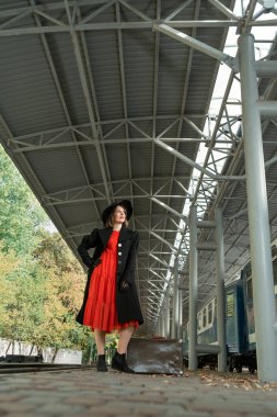 Kırmızı elbiseli, siyah paltolu ve şapkalı orta yaşlı aristokrat kadın tren istasyonunda bekliyor..