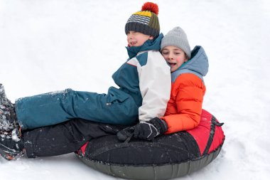 Erkekler şişme kızakla yamaçtan aşağı kayıyor. Kış kıyafetleri ve örgü şapkalı çocuklar kışın dışarıda eğlenirler.
