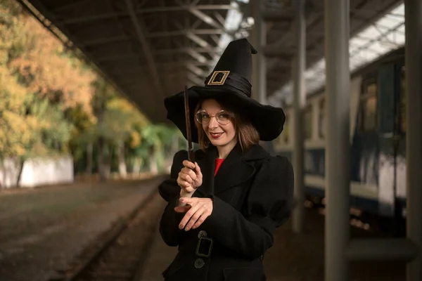 魔女の姿をした少女は謎の笑みを浮かべて 鉄道駅で魔法の杖を手にしている 衣装の魔術師 — ストック写真