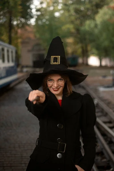 若い女性は 手に魔法の杖を持つ魔女の帽子 黒のコートや眼鏡でそりと服を着て笑顔 笑顔の女の子 — ストック写真