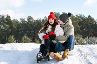 Genç bir adam ve bir kız oturur ve ormanın ve mavi gökyüzünün arka planında karın üzerinde kucaklaşırlar. Dışarıda bir çift..