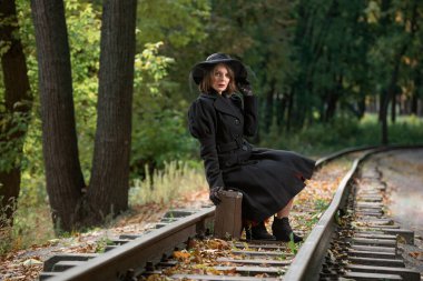 Klasik siyah paltolu ve şapkalı genç bir kadın ormandaki tren raylarında eski bir valizin üzerinde oturuyor. Klasik stil