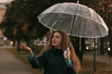 Kahverengi saçlı kız sonbahar parkında yürüyor. Yağmur yağıp yağmadığını görmek için şeffaf şemsiyenin altında duran genç kadının portresi.