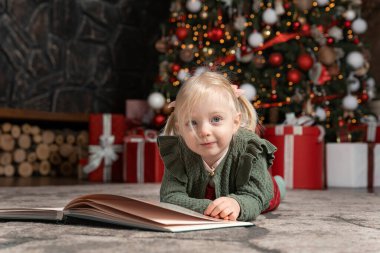 Noel ağacı arka planında büyük bir kitabın yanında yerde yatan tatlı sarışın kızın portresi. Çocuklar ve yeni yıl havası