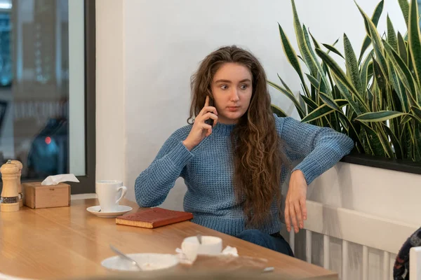 Teenager is talking on phone in cafe. Young girl with long brown hair sits in cafe with cup of tea or coffee and notebook.