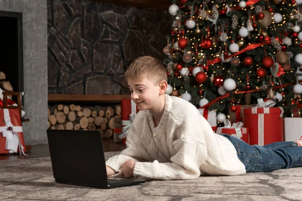 Boy with laptop near Christmas tree. Child received laptop as gift for New Year. Teenage watched Christmas movies