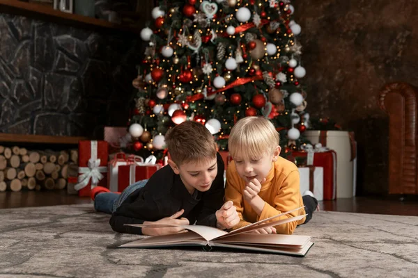 Two boys read book lying on floor under Christmas tree. Brothers or friends schoolboy read Christmas story or fairy tales.