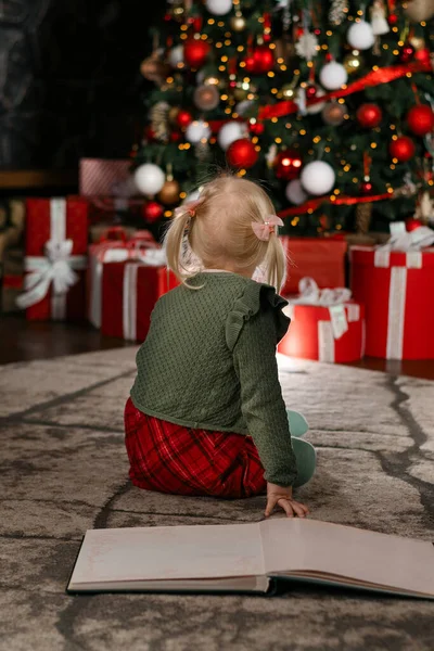 Little girl sits on the floor of row with large book and looks at beautiful Christmas tree. Back view. Vertical frame