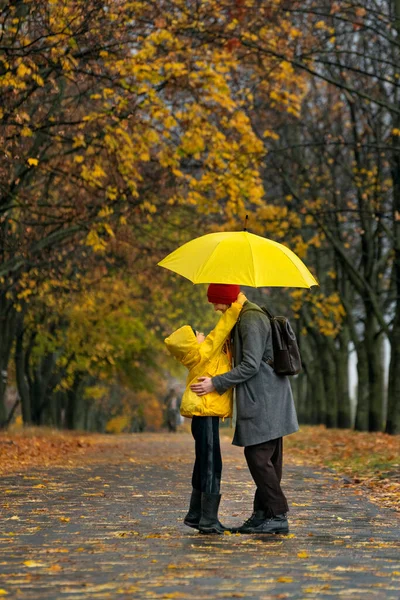Mom and child are walking in autumn park under large yellow umbrella. Vertical frame.