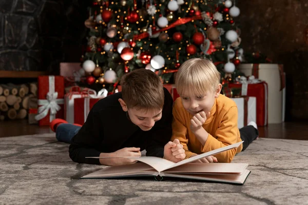 Two friends or brothers read book lying on floor on Christmas tree background. Children read Christmas story or fairy tales.