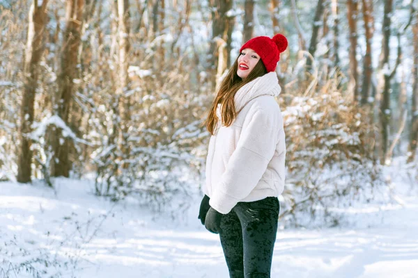 stock image Young beautiful girl in a red hat stands in a snow-covered winter sunny park. Outdoor recreation.