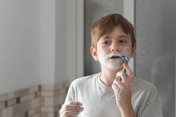 Schoolboy with shaving foam on face imitates shaving beard like his father. Boy in bathroom