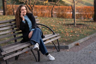Parkta bankta oturan, telefonla konuşan ve gülümseyen genç bir kadın. Genç kadın akıllı telefon kullanıyor..