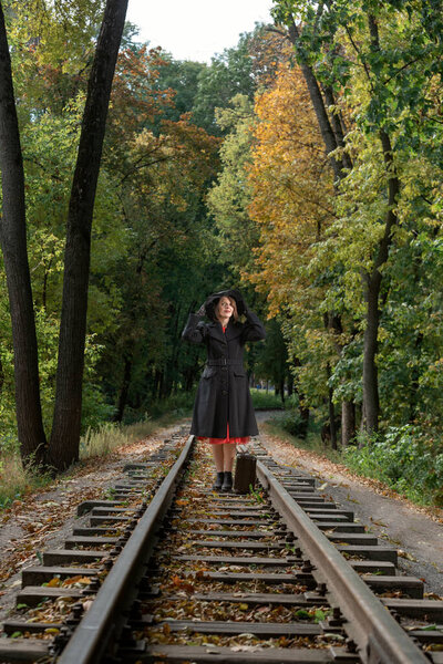 Woman in autumn forest in retro clothes black coat and broad-brim hat stands with old valise on rails. Woman dressed in 40s style.