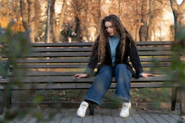 Düşünceli, uzun saçlı genç bir kadın sonbahar parkında bankta oturuyor ve sıkıcı. Sonbahar ılık günü