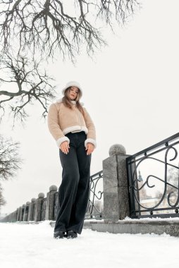 Siyah kot pantolon ve bej renkli koyun derisi ceketli, kar kaplı parkta şık bir kız. Dikey çerçeve.