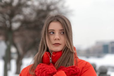 Kırmızı ceketli güzel bir kızın yakın plan portresi. Kırmızı örgü eldivenlerle elini ısıtıyor. Kışın açık havada, kalın giysiler içinde genç bir kadın..